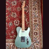 Světle zelená elektrická kytara typu Telecaster s javorovým hnedem. Na hlavě je viditelné logo Fender. Kytara má bílý pickguard a chromované kovové č…