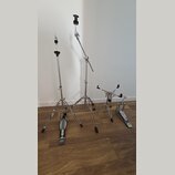 Sada chromovaných kovových stojanů a pedálů. V popředí jsou dva stojany s trojnožkou a jeden hi-hat stojan, vpravo je umístěn basový pedál. Pedály ma…