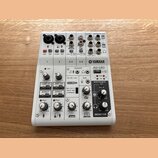 Bílá mixovací konzole značky YAMAHA, model AG06 MIXING CONSOLE. Zařízení obsahuje různé vstupy a výstupy, včetně konektorů XLR, jack 6,35 mm a USB. N…