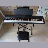 Černý digitální piano s klávesnicí, umístěné na černém X-stojanu s přiloženou černé polstrovanou židličkou. Na horní liště nástroje je uprostřed bílý…