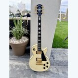 Elektrická kytara s krémovým povrchem korpusu a hmatníkem z tmavého dřeva s bílými blokovými vložkami. Na hlavě je zlatý nápis „Gibson“ a modrý ornam…