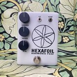 Světle šedý efekt pedál s nápisy HEXAFOIL a HARMONIC FUZZ. Na levé straně jsou tři černé oválné ovladače s popisky HARMONICS, DISTRESS a BLEND. V hor…