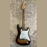 Elektrická kytara s trupem v barevnosti sunburst a bílým pickguardem. Na hlavě hmatníku je svisle napsaný nápis SKYLARK. Nástroj má tři snímače typu…