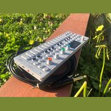 Bílý syntezátor OP-1 s klávesovou hrou a ovládacími knoflíky v různých barvách (modrý, zelený, oranžový). Na levé straně je viditelný nápis OP-1. Zař…