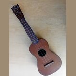 Hnědý dřevěný ukulele s tmavým hmatníkem. Nástroj vykazuje známky opotřebení, jsou vidět škrábance a nečistoty na povrchu těla. Na hlavě jsou struny…
