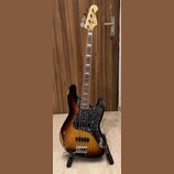 Elektrická basová kytara s trupem v barevnosti sunburst a černým pickguardem s texturou. Na hlavě hmatníku je nápis Fender Jazz Bass. Nástroj vykazuj…