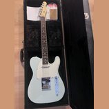 Světle modrá elektrická kytara typu Telecaster v černém tvrdém kufru. Na hlavě je viditelné logo Fender. Kytara má bílý pickguard, chromovaný most a…