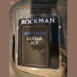 Kytarový procesor v černém obalu, který je umístěn v průhledném plastovém obalu. Na přední straně je bílý a modrý nápis ROCKMAN GUITAR ACE by Tom Sch…