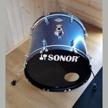 Modrý basový buben s černým předním koženem. Na koženě je bílý nápis SONOR s doprovodným logem. Na korpusu je vidět kovový úchyt a malý oválný štítek…