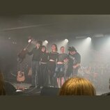 Fotografie zachycuje pět osob na pódiu v koncertním sále. Vlevo stojí muž s akustickou kytarou v rukou, vedle něj stojí čtyři další lidé. V pozadí je…