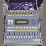 Šedý míchací pult s nápisem YAMAHA Programmable Mixer 01. Horní část obsahuje řadu přepínačů s žlutými papírovými štítky, uprostřed je umístěn LCD di…