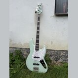 Světle zelená elektrická basová kytara s bílým perleťovým pickguardem. Na hlavě nástroje je viditelné logo "Squier". Tělo kytary má v dolní části čer…
