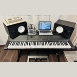 Digitální piano šedé barvy s nápisem ROLAND DIGITAL PIANO RD-1000, umístěné na černém stojanu s pedály. Na nástroji jsou umístěny dva černé studiové…