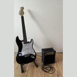 Černá elektrická kytara s bílým pickguardem a světle hnědým hmatníkem, umístěná na černém stojanu. Na hlavě kytary je viditelné logo Fender. Vedle ky…