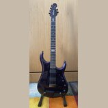 Tmavě fialová elektrická kytara s černým hmatníkem a dvěma humbuckery. Na hlavě je bílý nápis MUSIC MAN. Nástroj stojí na černém stojanu před dřevěno…