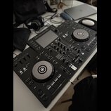 Černý DJ 컨트롤러 s dvěma jogovými koly a centrálním mixážním sekcí. Na levé straně je viditelný nápis XDJ-RR a v pravé části logo Pioneer DJ. Za přístro…