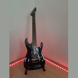 Černá elektrická kytara s lesklým povrchem a špičatou hlavou, umístěná na dřevěném stojanu. Na hlavě je viditelné logo LTD. Kytara má dva humbuckery,…