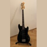 Černá elektrická basová kytara s hnědým hmatníkem a světlou dřevěnou hlavou. Na hlavě je nápis Fender MUSICMASTER BASS a logo Fender. Tělo má černý p…