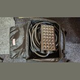 Černý rozpojitý kabel s konektory v kovovém boxu, umístěný v černé přepravní tašce. Na panelu jsou vidět početné konektory XLR a nápisy jako M1, M2,…