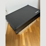 Černý transportní kufr s texturovaným povrchem a kovovými hranami. V pravém horním rohu je bílé logo MAGMA. Kufr stojí na dvou kartonových krabicích…