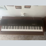 Černý digitální klavír umístěný na hnědém dřevěném stole. Na pravé straně je bílý nápis YAMAHA a vedle něj modelové označení pf15. Nástroj má standar…