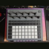 Šedý sampler Novation Circuit Rhythm s bílými podsvícenými pady v mřížce 4x8. V horní části jsou umístěny otočné ovladače s popisky Master Volume, Tu…