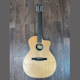 Akustická kytara s vyřezem (cutaway) a přírodní barvou dřeva. Na hlavě je viditelné logo "Taylor". Nástroj vykazuje čistý povrch bez viditelných pošk…