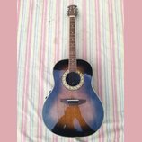 Akustická kytara s cutawayem a tmavým, přechodovým lakováním (sunburst). Kolem otvoru rezonátoru je viditelná ozdobná rozeta se zlatými vzory. Na hla…