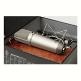 Stříbrný kondenzátorový mikrofon s kovovou síťkou v horní části, umístěný v černé krabici na dřevěném podkladu. Na těle mikrofonu je viditelné logo z…
