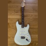 Bílá elektrická kytara s bílým pickguardem a hnedým hmatníkem. Na hlavě je viditelné logo Fender Stratocaster. Nástroj má tři snímače s bílými krytka…