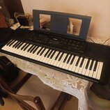 Černý digitální klavír s bílými a černými klávesami, umístěný na stole s bílým ubrusem. Na ovládacím panelu je v centru bílé logo YAMAHA a displej. N…