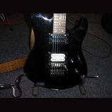 Černá elektrická kytara s třemi snímači (dvě humbuckery a jeden single-coil). Kytara má hnědý dřevěný hриф a kovový tremolo systém s páčkou. Na těle…
