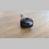 Černý plastový knoflík s nápisem Roland. Je připevněn na kovovou osa a položen na dřevěném povrchu. Povrch plastu vykazuje drobné škrábance.