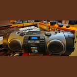 Šedý přenosný radiomagnetofon značky JVC. Na předním panelu je digitální displej, ovládací tlačítka a nápis POWERED BOOMBOX CD SYSTEM RV-KN401. Ve sp…