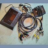 Černý elektronický přístroj s nápisy "CYMBIC AUDIO" a "Live Pipe LP 16". Součástí je černý napájecí adaptér, černý kabel svázaný na hranatém podložko…