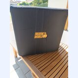 Černý textilní kryt na hudební zařízení, umístěný na dřevěném stole. Uprostřed je žluté logo s nápisem MARK WORLD a stylizovanou postavou. Povrch lát…