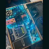 Dřevěný kufr s pedalboardem, který je podsvícen modrými LED pásky. Obsahuje efektové pedály a procesor s nápisy „BOSS“, „MB-800“ a „MAH SUP“. V kufrě…