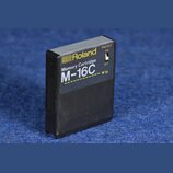 Černá paměťová karta z plastu s žlutými nápisy. Na přední straně je logo a název Roland, text Memory Cartridge a označení modelu M-16C. Na pravém bok…