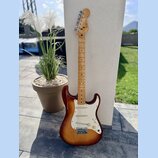 Elektrická kytara s trupem v barevnosti sunburst a hnedým hmatníkem. Na hlavě je viditelné logo Fender. Kytara má bílý pickguard, tři snímače a kovov…