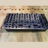 Stříbrný DJ mixér s černými ovladači a posuvníky. Na levém boku je nápis VMX 1000 a na pravém boku logo BEHRINGER. Uprostřed spodní části je nápis XP…