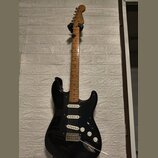 Černá elektrická kytara s hnědým dřevěným hmatníkem a bílými snímači. Na hlavě nástroje je viditelné logo Squier. Kytara má bílé ovládací knoflíky a…