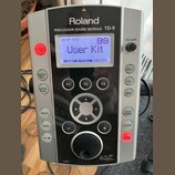 Stříbrno-černý perkusní modul značky Roland s nápisem PERCUSSION SOUND MODULE TD-9. Displej zobrazuje text DRUM KIT 99 User Kit a spodní menu INST, F…