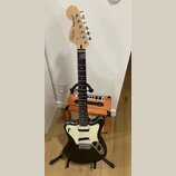 Elektrická kytara v černé barvě s bílým perleťovým pickguardem, umístěná na černém stojanu. Na hlavě hmatníku je nápis Squier by Fender a modelové oz…