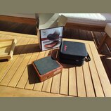 Na dřevěném stole je umístěn Stompbox v dřevěném provedení s černou horní plochou a nápisem "Shadow". Vedle něj je černý textilní přepravní pouzdro s…