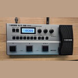 Stříbrný basový efektový procesor s černým výrazným pedálem vpravo. Na horní části je nápis BOSS GT-1B BASS EFFECTS PROCESSOR a na displeji logo BOSS…