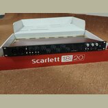 Černý rackový zvukový rozhraní umístěný v původním červenobílém obalu. Na přední straně zařízení je nápis Scarlett 18i20 a logo Focusrite. Přístroj o…