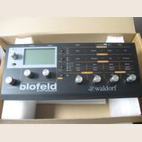 Šedý desktopový syntetizátor s nápisy blofeld SYNTHESIZER a waldorf. Přístroj má displej, několik ovládacích knoflíků a řadu tlačítek s popisky jako…