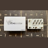 Bílá krabice a efekt pedál s bílým povrchem. Na obou je černý nápis ECHO SYSTEM DUAL ENGINE DELAY a logo Empress. Pedál obsahuje několik ovládacích k…