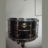 Černý malý snare drum s chromovaným kováním. Na koruse je umístěn oválný štítek s nápisy „pawpaw“, „BLACK MAGIC“ a „SNARE DRUM LIMITED“. Povrch nástr…