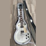 Bílá elektrická kytara s tvarem singlecut v černém etuši s bílou vnitřní výplní. Na hlavě je viditelné logo Gibson. Kytara má dva humbuckery, světle…